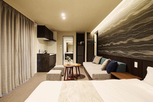 A modern hotel room with a bed, sofa, and a small table. There is a mirror, a wardrobe, and a view of the outside.