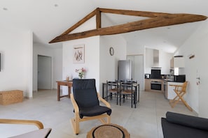 A modern living room with a dining area, kitchen, and a loft bedroom with exposed wooden beams.