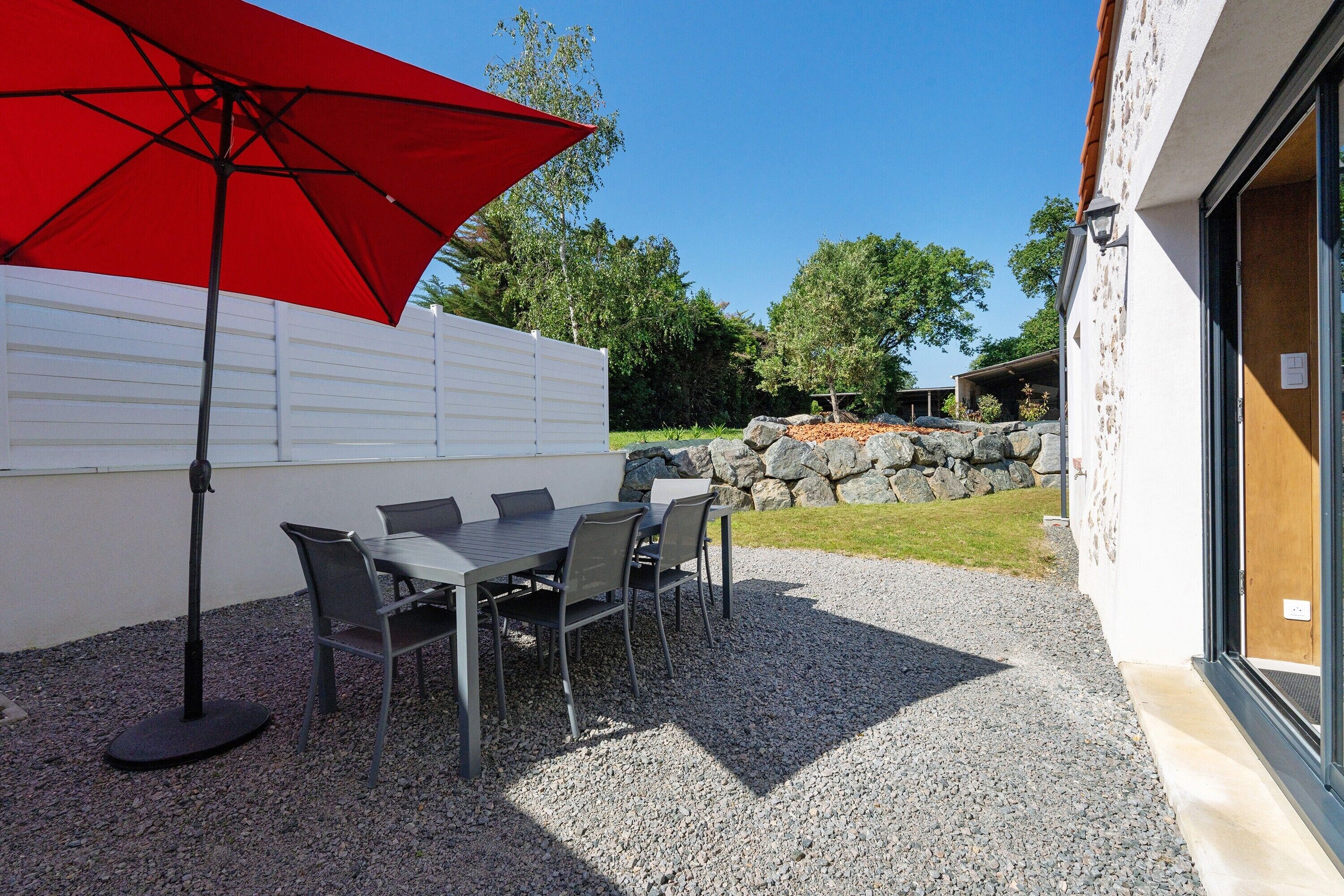Une terrasse extérieure avec une table et des chaises, un parasol rouge et un mur blanc.