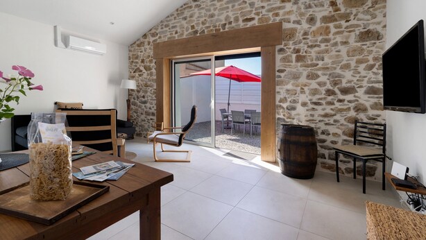 A spacious room with a stone wall, a wooden dining table, a comfortable chair, a TV mounted on the wall, and a sliding glass door leading to an outdoor patio area. - Holiday Home 'Gîte L'atelier - 4 Personnes' with Private Terrace, Wi-Fi and Air Conditioning (Frossay)