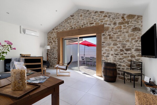 A spacious room with a stone wall, a wooden dining table, a comfortable chair, a TV mounted on the wall, and a sliding glass door leading to an outdoor patio area.
