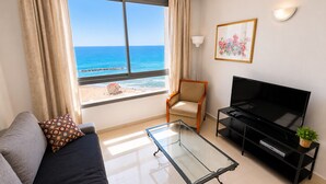 A living room with a sofa, a glass coffee table, a TV on a black stand, a chair, and a large window with a view of the ocean. - Azure Beach Apartments (Haifa)