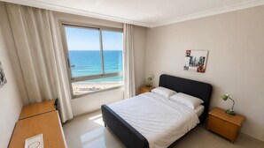 A hotel room with a bed, a desk, a nightstand, a lamp, a picture on the wall, and a view of the ocean. - Azure Beach Apartments (Haifa)