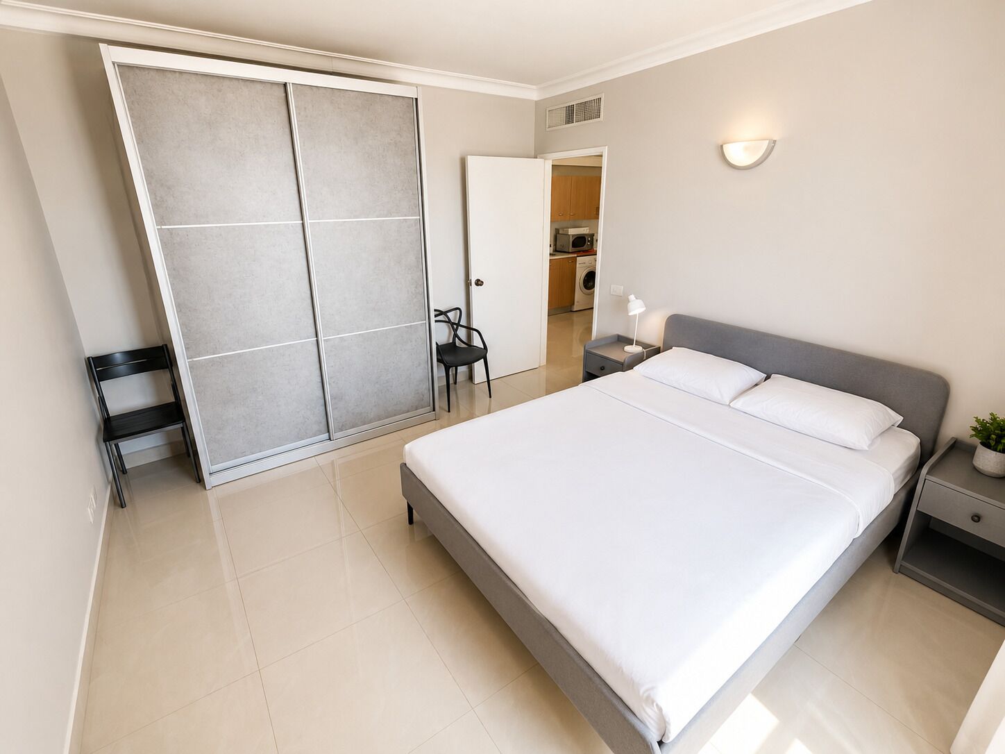 Une chambre à coucher comprenant un lit, une table de chevet, une lampe, une chaise, une armoire et donnant sur une cuisine.