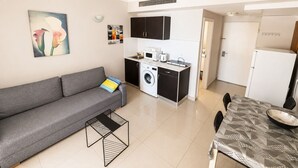 A compact living space with a sofa, a dining table, a washing machine, and a kitchenette. - Azure Beach Apartments (Haifa)