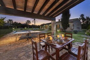 Ein Essbereich im Freien mit einem Steintisch und Stühlen, einem Pool und einem Steinhaus im Hintergrund.