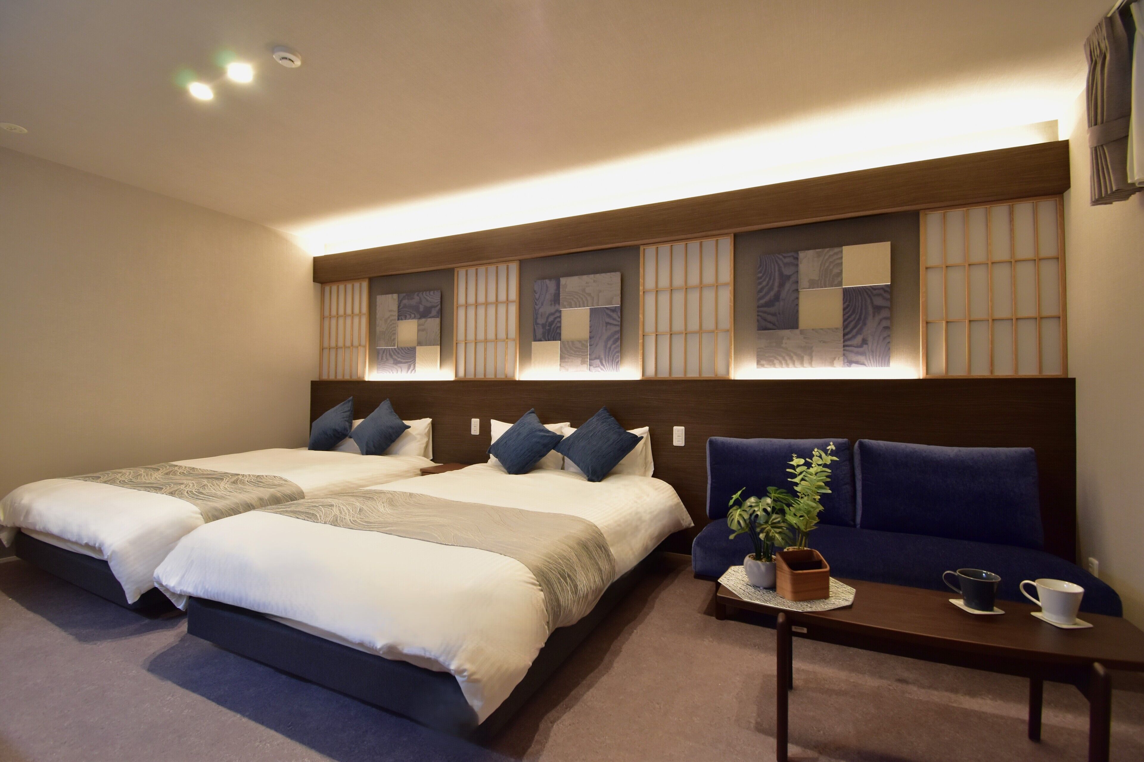 A hotel room with two beds, a sofa, a small table with a plant, and a wall with decorative panels.