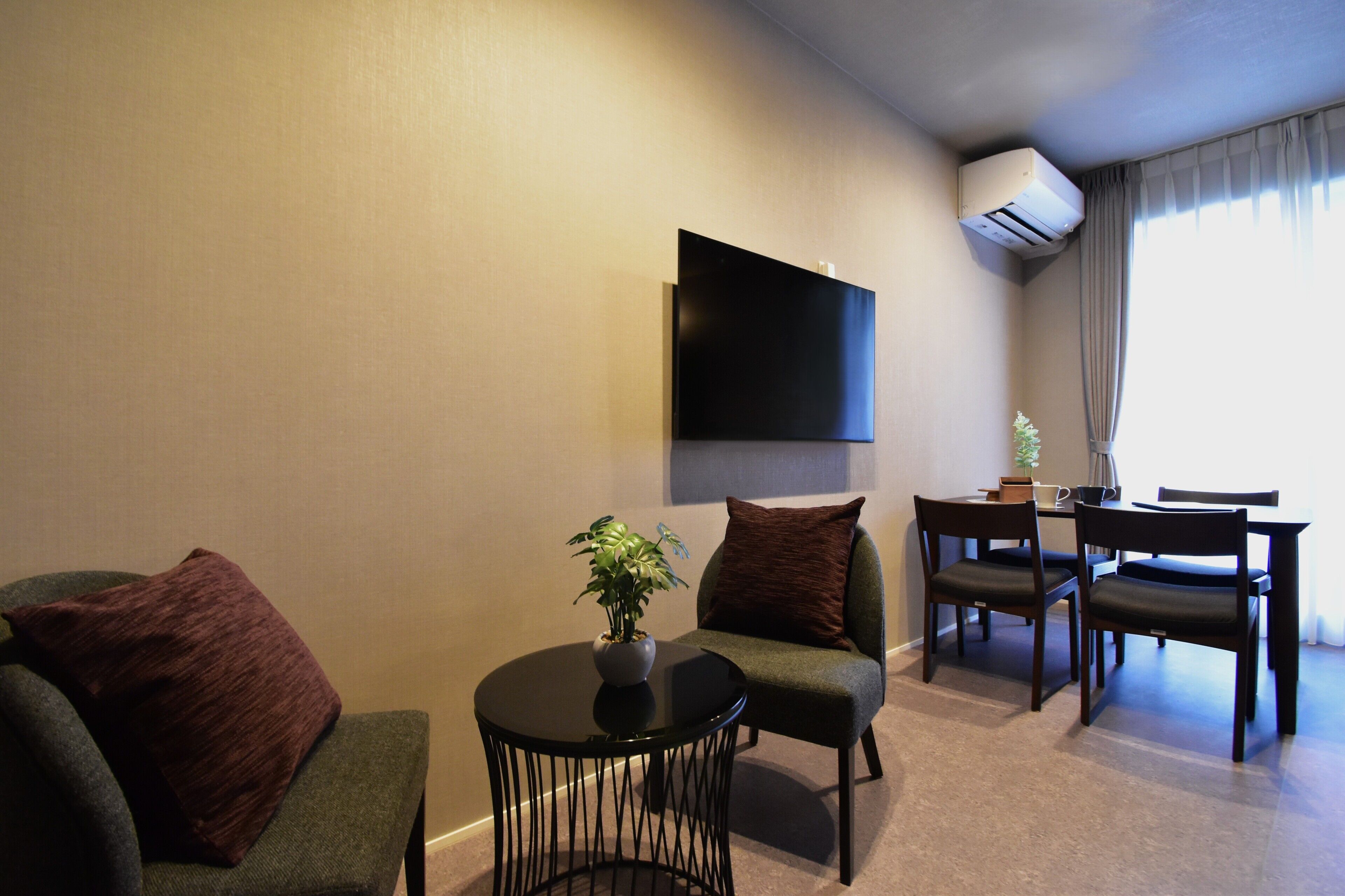 A living room with a sofa, chairs, a small table with a plant, a flat-screen TV mounted on the wall, a dining table with chairs, and an air conditioning unit.