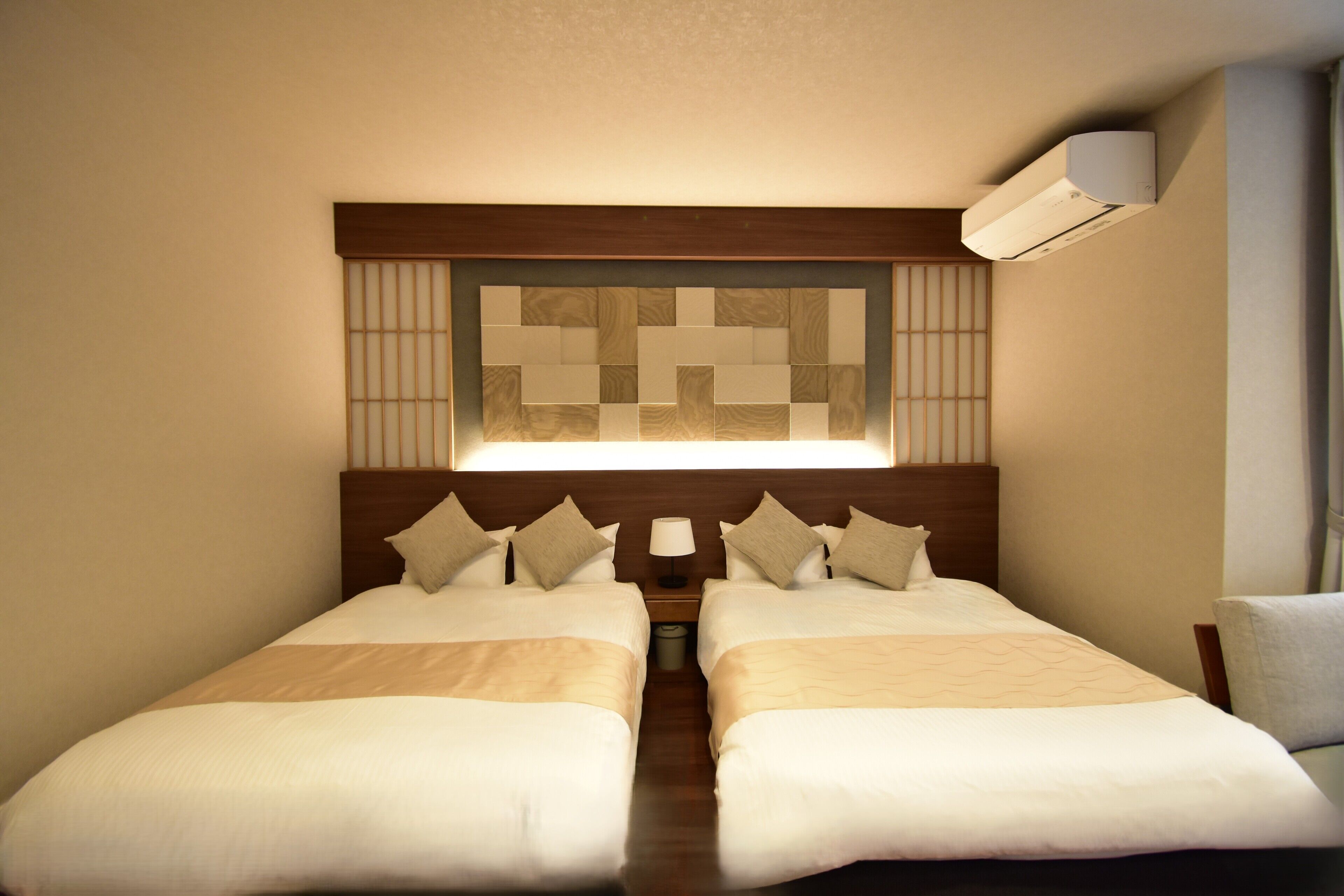 A hotel room with two beds, a wooden headboard, a wall-mounted air conditioner, and a nightstand with a lamp.