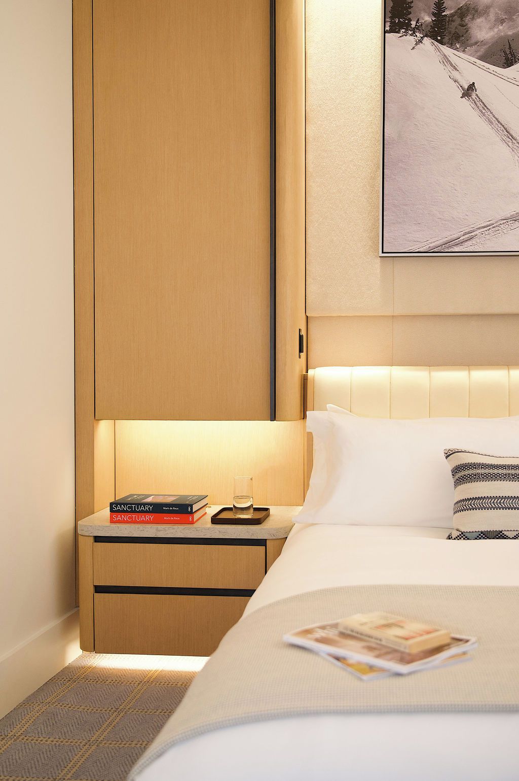 A hotel room with a bed, a nightstand with books and a glass, a wooden wardrobe, and a framed picture on the wall.