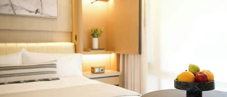 A hotel room with a neatly made bed, a bedside table with a lamp, and a fruit bowl on a side table.