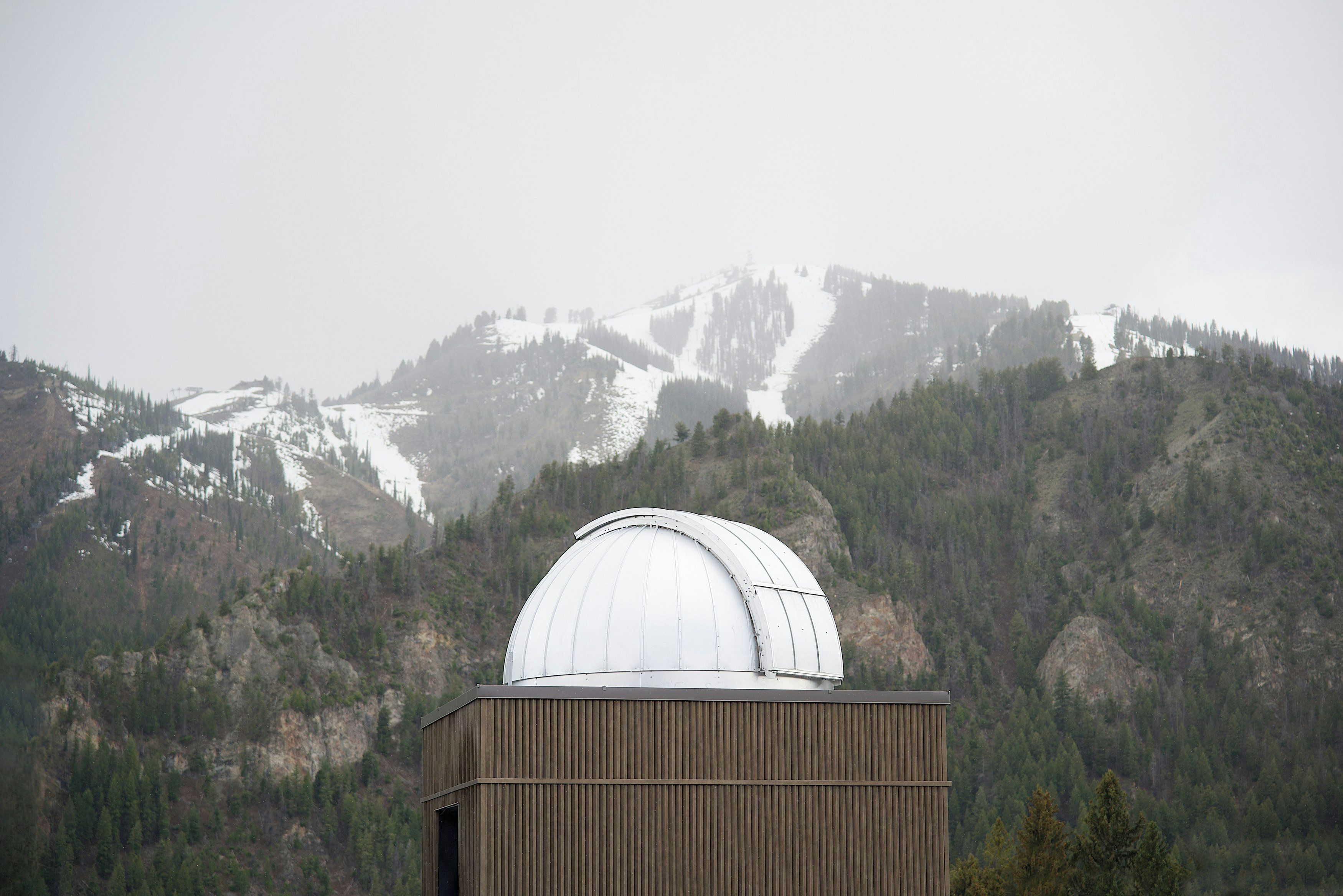 A dome-shaped observatory with a corrugated metal exterior, set against a backdrop of snow-capped mountains and dense forest.