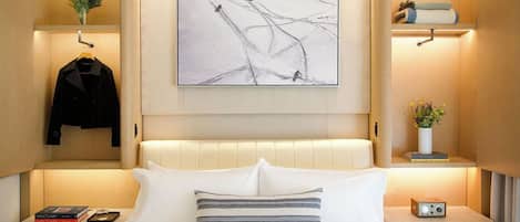 A hotel room with a bed, a nightstand, a bedside table, a wall-mounted painting, and a shelf with a jacket and a plant.