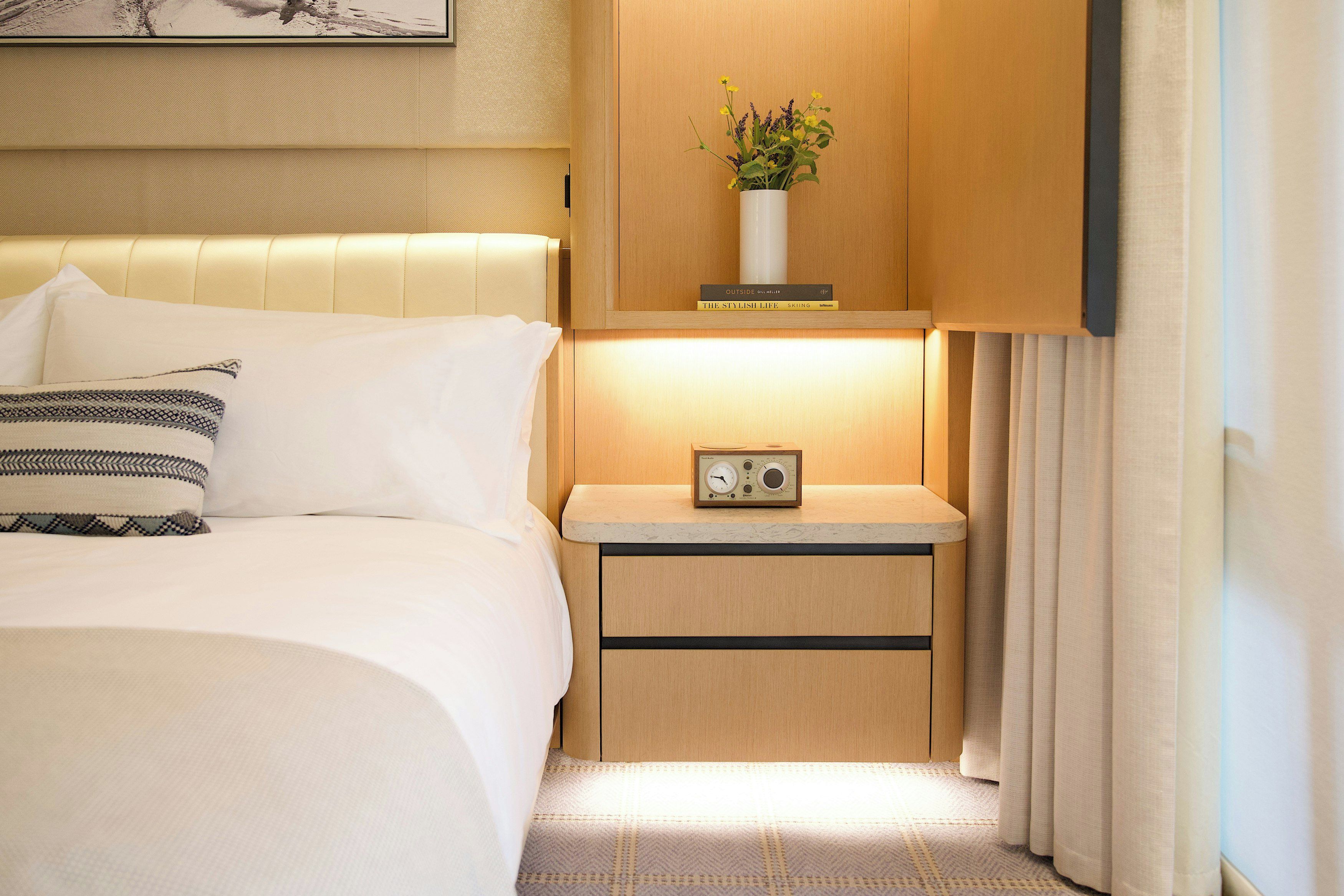 A hotel room with a bed, bedside table, radio, and a small plant on the nightstand.