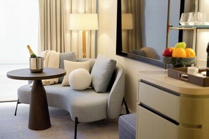 Una habitación de hotel moderna con un sofá gris, una mesa redonda con una botella de champán, un espejo y un frutero sobre la encimera.