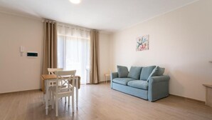 A living room with a light blue sofa, a dining table with chairs, a window with curtains, and a wall-mounted light fixture. - Agriturismo Cavalieri (Civita Castellana)
