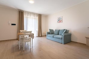 Una sala de estar con un sofá azul claro, una mesa de comedor con sillas, una ventana con cortinas y una lámpara fijada a la pared.