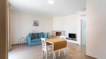 Una sala de estar con un sofá azul, una mesa de comedor de madera con sillas, una pequeña mesita auxiliar blanca, una chimenea y un televisor.