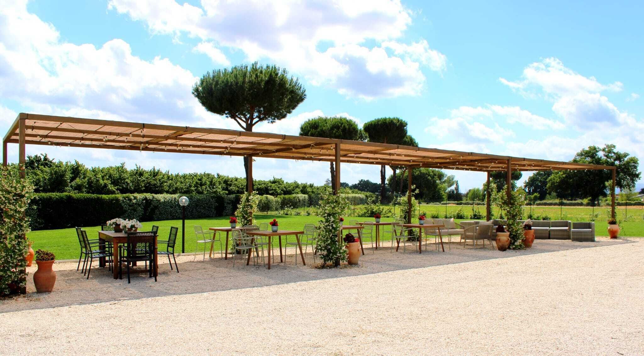 Un patio exterior con mesas y sillas, una pérgola y un jardín cuidado.
