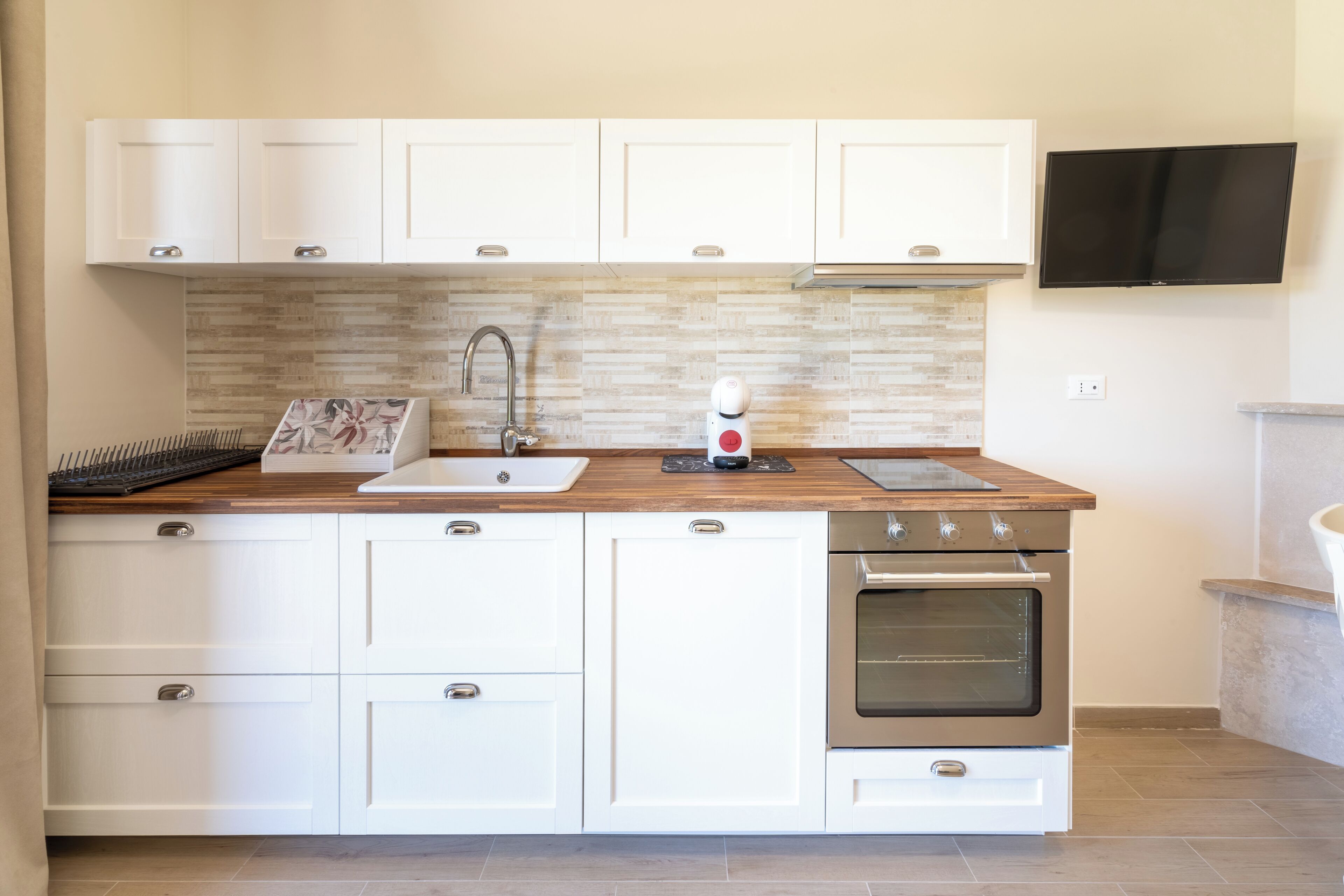 Een moderne keuken met witte kasten, een houten aanrecht, een inbouwoven en een aan de muur bevestigde televisie.