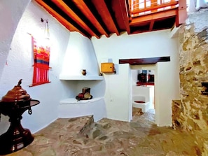 Un couloir étroit aux murs de pierre, avec un plafond blanc et une poutre en bois au-dessus. On aperçoit une petite étagère sur laquelle se trouvent une tasse et un pot, et une porte donne accès à une autre pièce.
