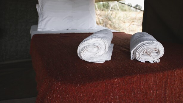 Una cama con sábanas blancas, una colcha roja y una vista de un paisaje seco a través de una ventana.
