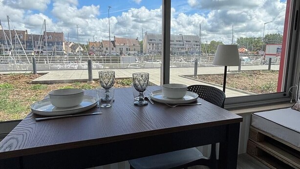 Een eethoek met een tafel voor twee personen, grote ramen en uitzicht op gebouwen en een parkeerplaats.
