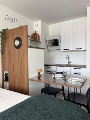 A modern kitchen with a wooden door, a clock, a TV, and a dining area with a table set for two. - Voile du cap, studio with loggia overlooking the port (Courseulles-sur-Mer)