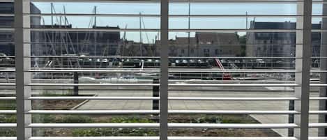 A window with horizontal blinds, a vase with a plant, and a view of an airport with buildings and cranes.
