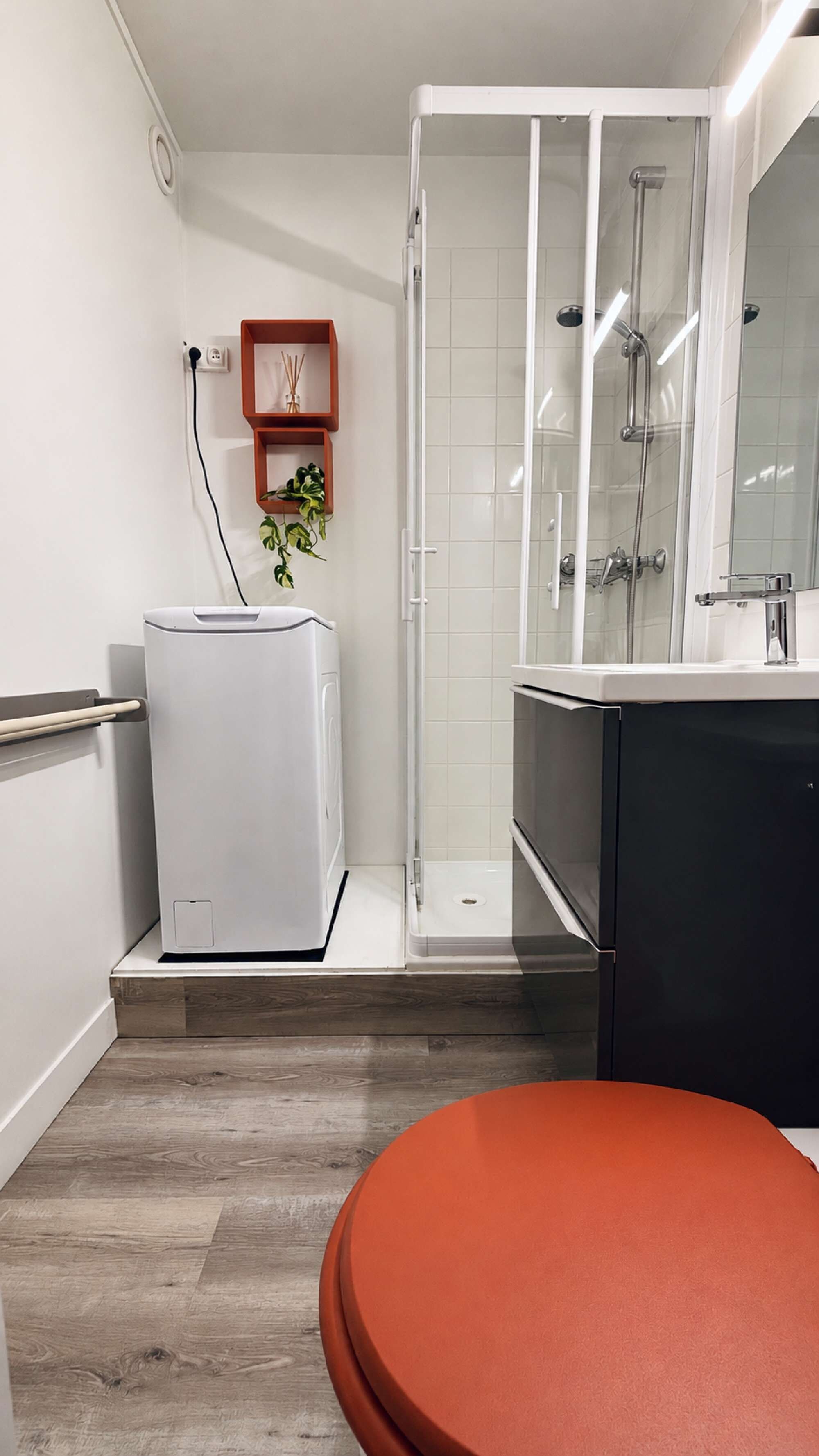 Een moderne badkamer met een glazen douchecabine, een witte wasmachine, een zwart wastafel en een houten vloer.