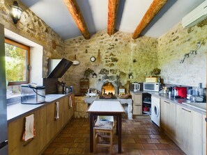 A rustic kitchen with stone walls, wooden beams, a stone fireplace, a dining table, and modern appliances. - Charming house in Provence with outbuilding - Swimming pool - 15 people (Aurons)