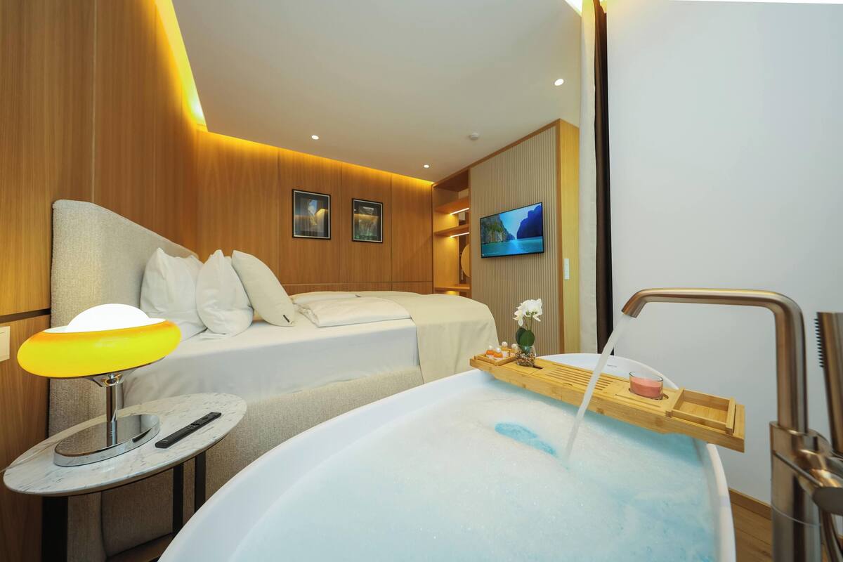 Una habitación de hotel con cama, una mesita con lámpara, una bañera con bandeja y un televisor en la pared.