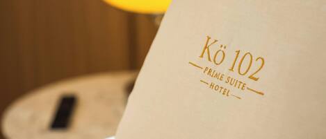 A pillow with 'Kö 102 Prime Suite Hotel' text, a bedside lamp, and a wooden nightstand.