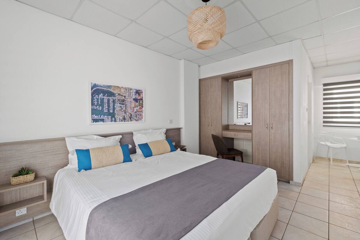 A modern hotel room with a large bed, a desk, a chair, a hanging light fixture, and a cityscape artwork on the wall.