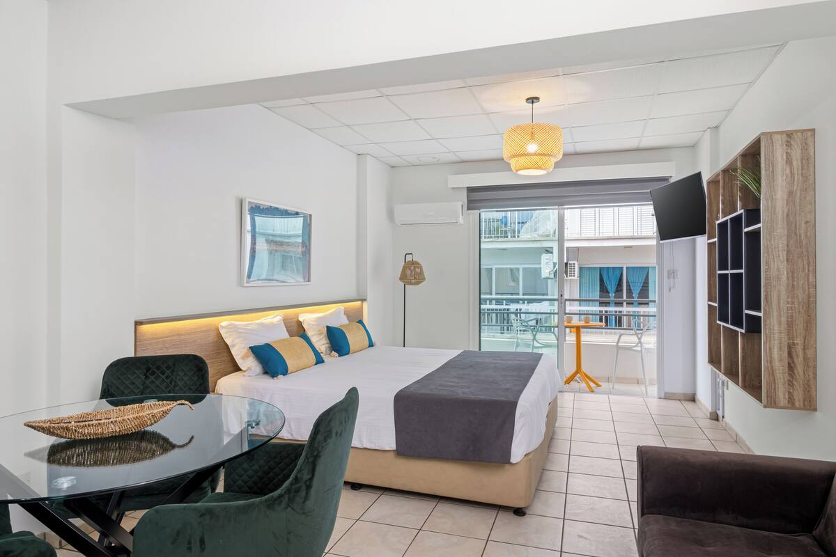 A modern hotel room with a bed, a glass table, chairs, a TV, and a balcony with a view.