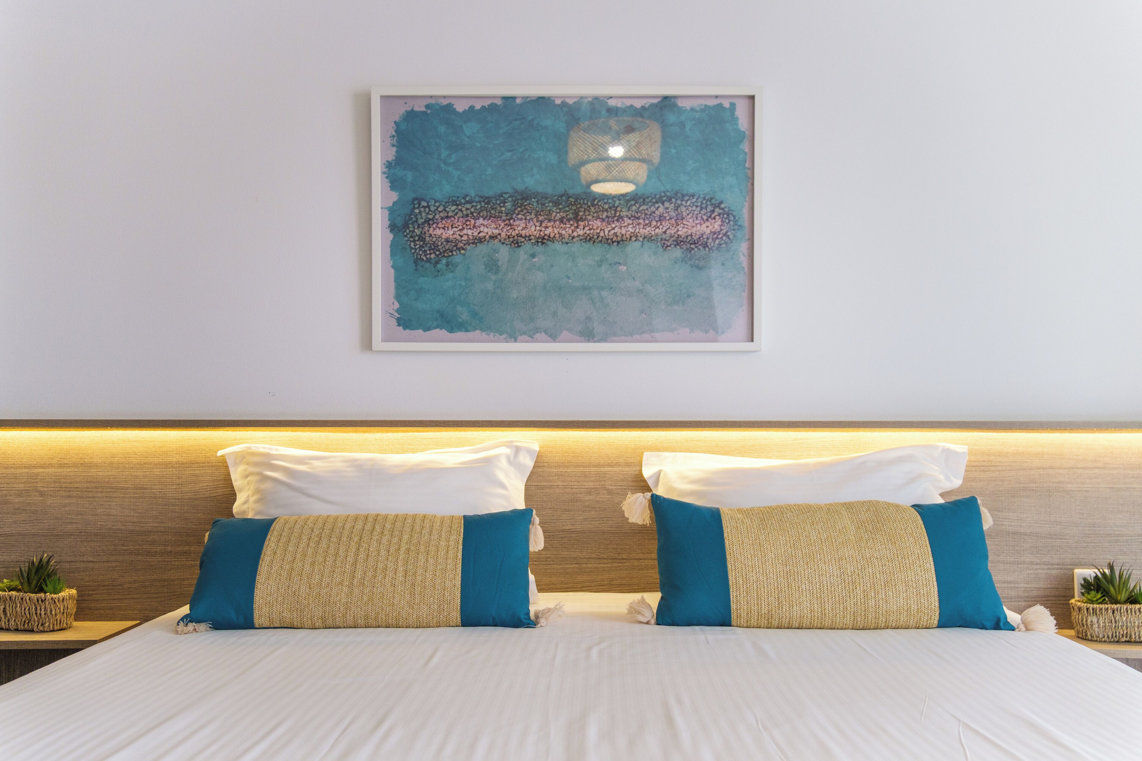 A neatly made bed with white and blue bedding, a headboard with a wooden finish, and a framed abstract painting above it.