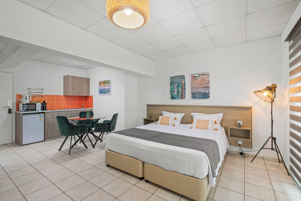 A modern hotel room with a bed, bedside tables, a desk with a chair, a kitchenette, and a dining area.