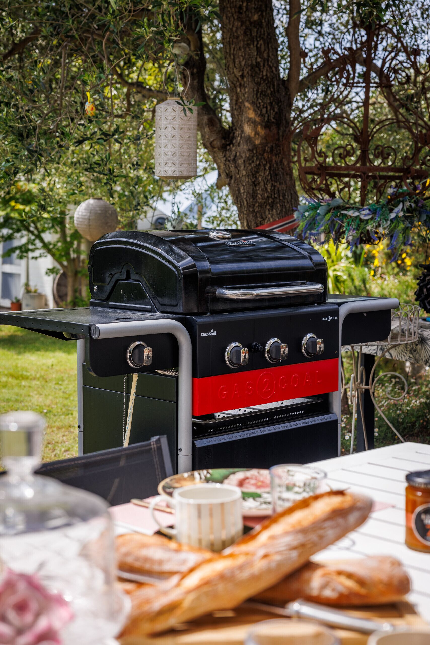 A barbecue grill with a gas supply, a table with bread and drinks, and a tree in the background.