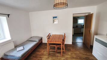 Ein Zimmer mit einem Holztisch, einem Einzelbett mit Kissen, einer Pendelleuchte und Blick zur Küche.