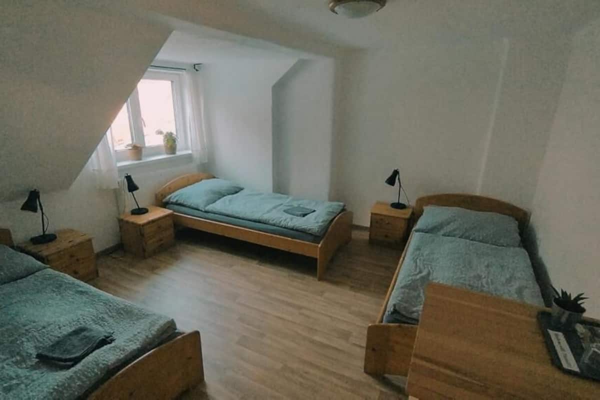 A dormitory room with three wooden bunk beds, a window with a view, and a small table with a lamp.