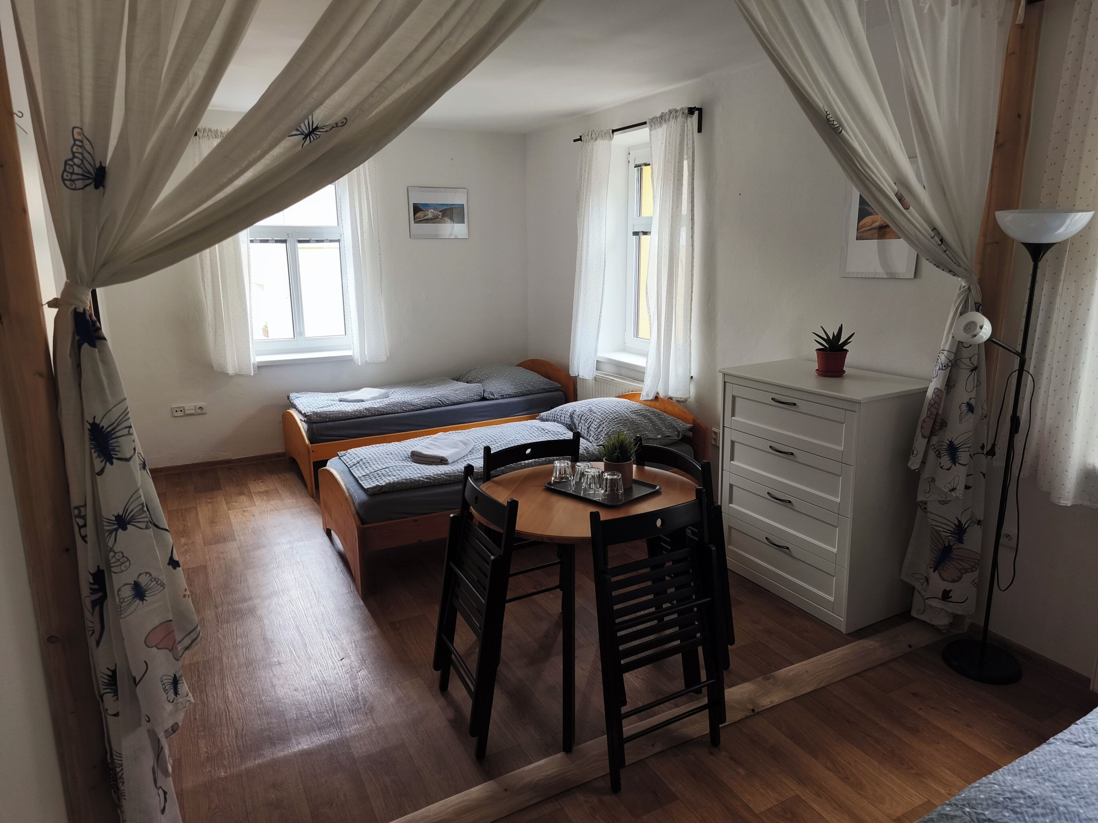 A dormitory room with two single beds, a wooden table with chairs, a white dresser, and a window with curtains.