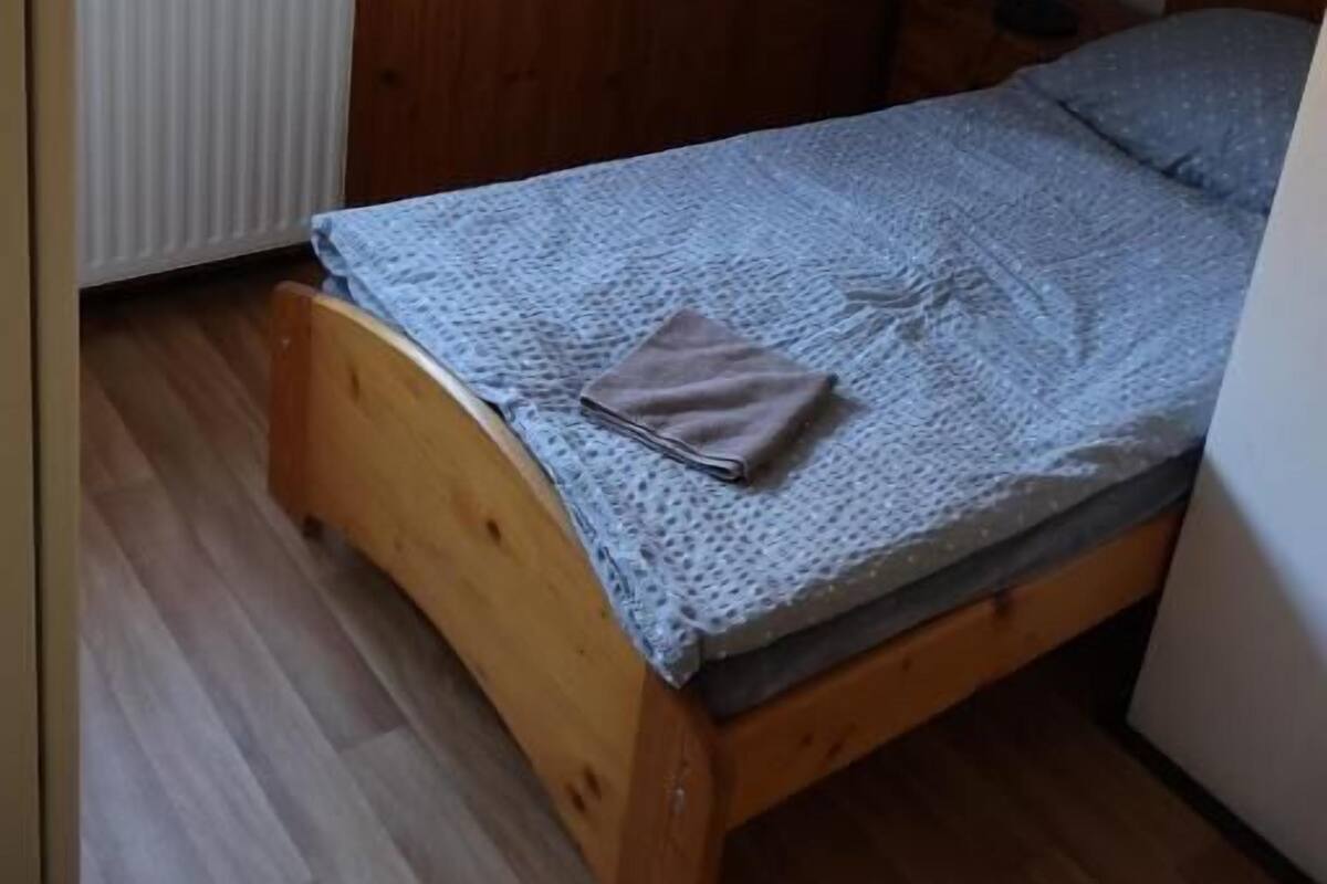 A small, wooden-framed room with a sloped ceiling, a single bed with a patterned blanket, a radiator, and a window.