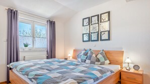 Ein Schlafzimmer mit Bett, Nachttischen, einem Fenster mit Vorhängen, einem Heizkörper und Wandbildern.