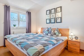 Ein Schlafzimmer mit Bett, Nachttischen, einem Fenster mit Vorhängen, einem Heizkörper und Wandbildern.