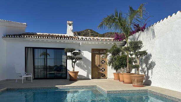 A poolside area with a clear pool, a white building with a tiled roof, and potted plants. - Villa La Casita Fuente del Baden 52 (Nerja)