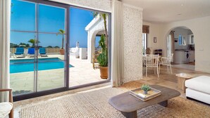 A spacious living area with a large sliding glass door leading to a pool, a dining area with a table and chairs, and a kitchen visible through an archway. - Villa La Casita Fuente del Baden 52 (Nerja)