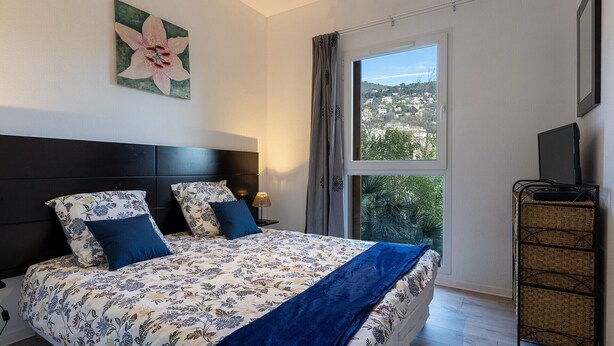A bedroom with a bed, a nightstand, a lamp, a TV, and a window with a view of a town. - Bright apartment for 4 persons with a mountain view (Grasse)