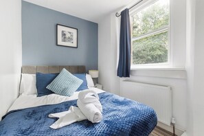 Ein ordentlich bezogenes Bett mit blauer Decke und Kissen, ein gerahmtes Bild an der Wand und ein Fenster mit Vorhängen.