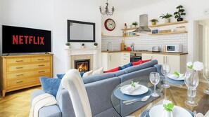 Ein modernes Wohnzimmer mit Kamin, einer Couch, einem Esstisch für zwei Personen und einer Küchenzeile mit weißen Schränken und einer Mikrowelle.
