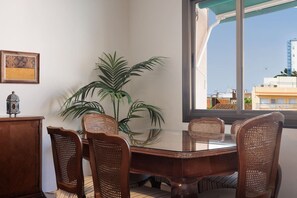 Un comedor con una mesa de vidrio, sillas de mimbre, una planta en maceta y vistas de edificios a través de una ventana.
